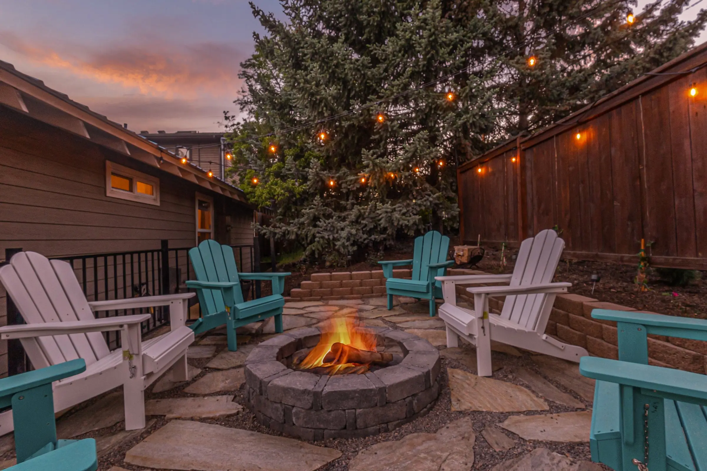 Backyard firepit glowing at dusk under string lights at Magnolia House vacation rental in Bend, Oregon, with teal Adirondack chairs circled around the flames